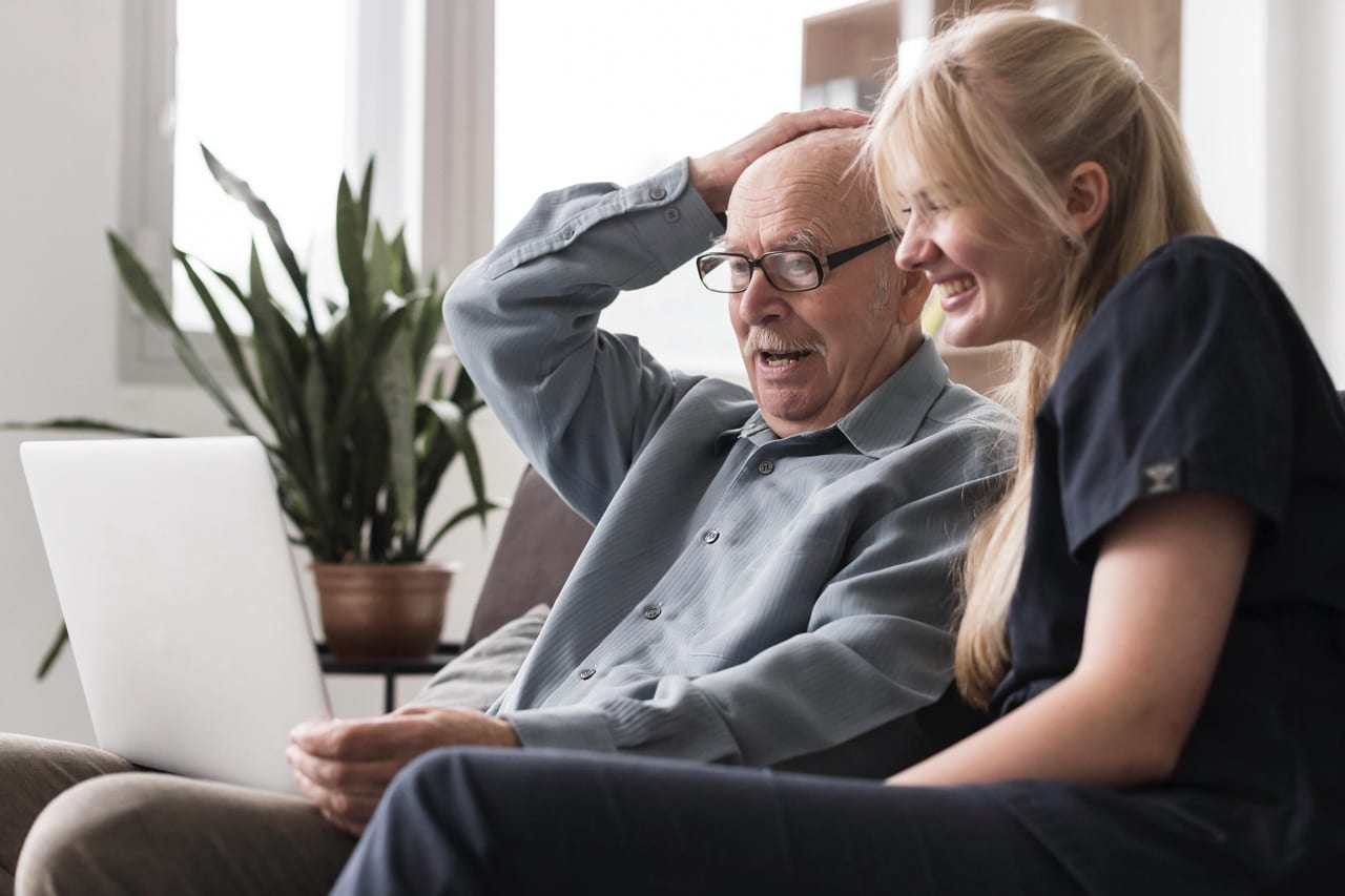Eine jüngere blonde Frau sitzt neben einem älteren Herrn auf der Couch. Sie schauen gemeinsam auf einen Laptop.