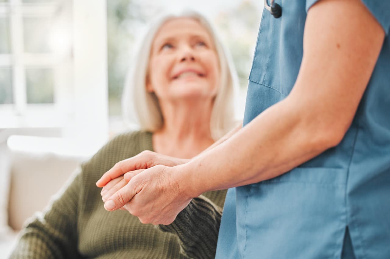 Eine Frau in blauem Hemd hält die Hand einer etwas älteren, lächelnden Dame.